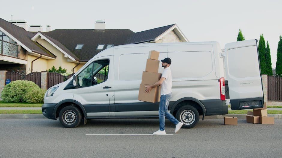A man dressed in a blue jacket and dark trousers is seen loading or unloading a white cargo van parked along a city street. The van's rear doors are open, revealing the empty interior with visible woo