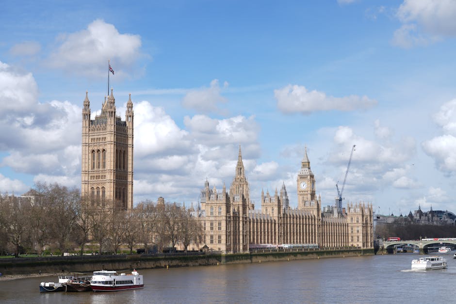 A daytime view of the Palace of Westminster in London, featuring the iconic Big Ben clock tower with its clock face visible, situated beside the River Thames. The historic Gothic-style building showca